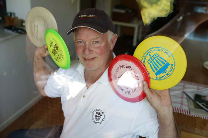 Ein älterer Mann mit schwarzem Basecap, weißem Shirt und mehreren bunten Frisbees