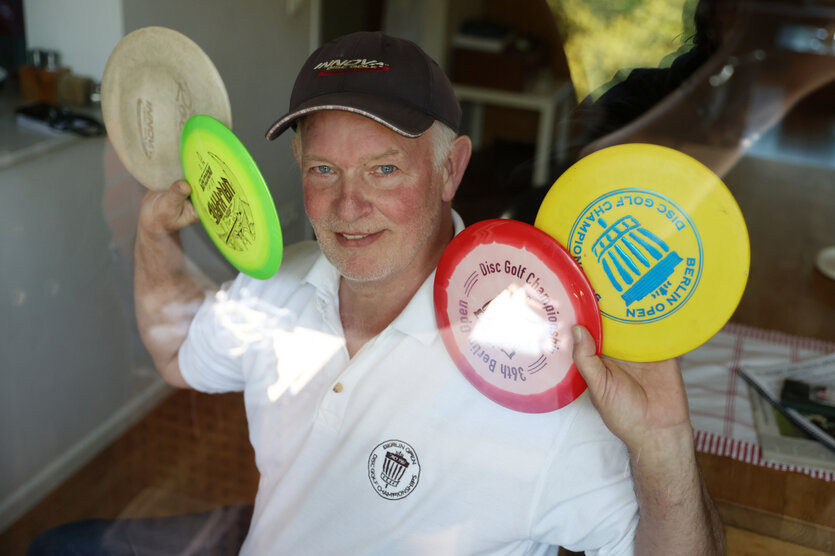 Ein älterer Mann mit schwarzem Basecap, weißem Shirt und mehreren bunten Frisbees