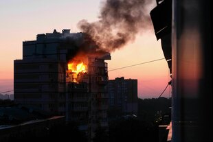 Eine Wohnung in einem mehrstöckigen Wohnhaus brennt nach einem russischen Drohnenangriff, der Block ist an sich noch in Nachtschwarz, das Feuer und der Sonnenaufgang heben sich jedoch bereits ab.