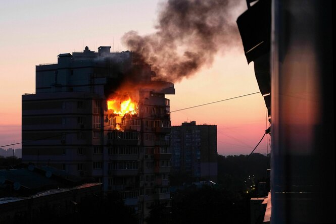 Eine Wohnung in einem mehrstöckigen Wohnhaus brennt nach einem russischen Drohnenangriff, der Block ist an sich noch in Nachtschwarz, das Feuer und der Sonnenaufgang heben sich jedoch bereits ab.