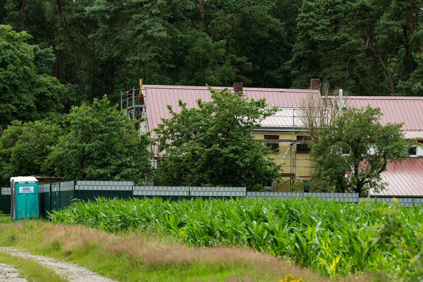 Zwei umzäunte Gebäude vor einem Wald. An den Fassaden sind Baugerüste aufgestellt.