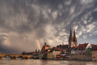 Sicht auf Regensburg, vom Fluss aus gesehen, Brücke und Kirche im Mittelgrund, Schwere Wolken im Himme und ein zarter Regenbogen