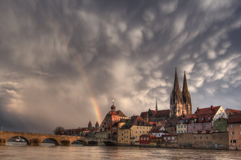Sicht auf Regensburg, vom Fluss aus gesehen, Brücke und Kirche im Mittelgrund, Schwere Wolken im Himme und ein zarter Regenbogen