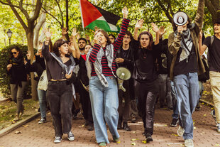 Protestierende mit erhobenen Händen laufen mit Kufiyas, Palästina-Flagge und Megafon auf die Kamera zu