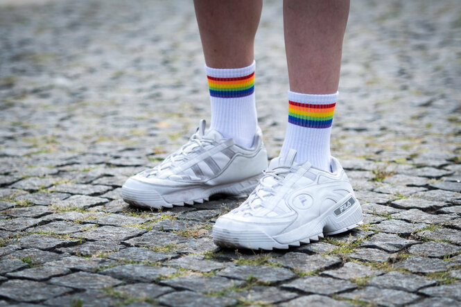 Detailaufnahme, Füße einer Person in weißen Turnschuhen und Socken, die Socken sind von der Regenbogen Flagge bekrönt