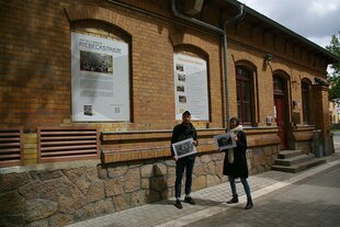 Zwei Menschen stehen vor der Gedenkstätte Riebeckstraße 63 in leipzig und halten historische Fotos in den Händen