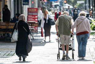 Passantinnen und Passanten gehen am Morgen durch eine Straße in Berlin-Moabit. Im Vordergrund sind eine Frau mit Kopftuch sowie ein älteres Paar mit Rollator zu sehen
