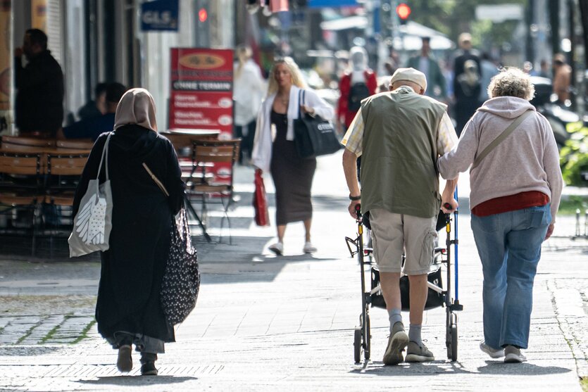 Passantinnen und Passanten gehen am Morgen durch eine Straße in Berlin-Moabit. Im Vordergrund sind eine Frau mit Kopftuch sowie ein älteres Paar mit Rollator zu sehen