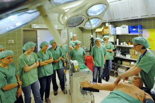 Ein Bild aus einem Operationssaal: Mehrere Personen in grünen OP-Kitteln stehen um einen Tisch mit einem Patientenmodell, das mit Schläuchen und Elektroden verbunden ist. Über dem Tisch hängt eine OP-Lampe. Im Hintergrund sind Regale mit medizinischen Ger