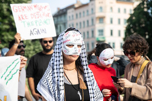 Eine Person mit palituch über dem Kopf und Maske im Gesicht steht auf der Straße, hinter ihr andere Menschen und ein Altbau