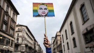 Jemansd hält ein SChild in die Höhe, darauf sieht man einen grell geschminkten Viktor Orban vor bunten Streifen in Regenbogenfarben