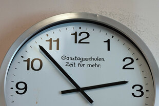 Eine Wanduhr zeigt sieben Minuten vor drei an, auf dem Uhrenblatt steht: 