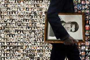 Ein Mann mit einem Porträtfoto geht an einer Fotowand vorbei, auf der Ermordete des iranischen Mullahregimes aufgereiht sind