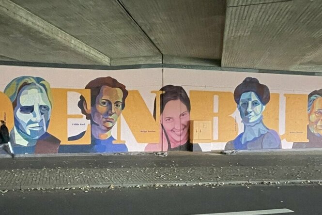 Eine Wandmalerei unter einer Brücke mit sechs farbigen Porträts von Personen, die zwischen großen gelben Buchstaben "DENKBU" angeordnet sind. Ein Mensch steht davor auf einer Straße.