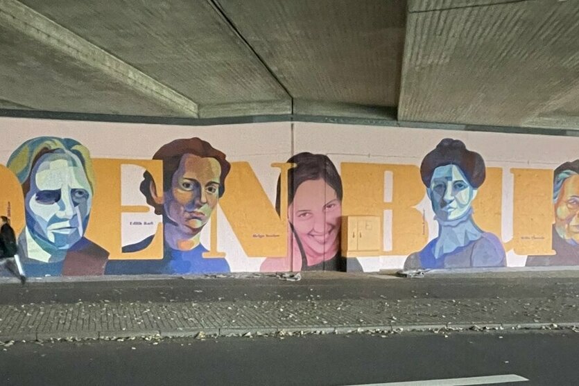 Eine Wandmalerei unter einer Brücke mit sechs farbigen Porträts von Personen, die zwischen großen gelben Buchstaben "DENKBU" angeordnet sind. Ein Mensch steht davor auf einer Straße.