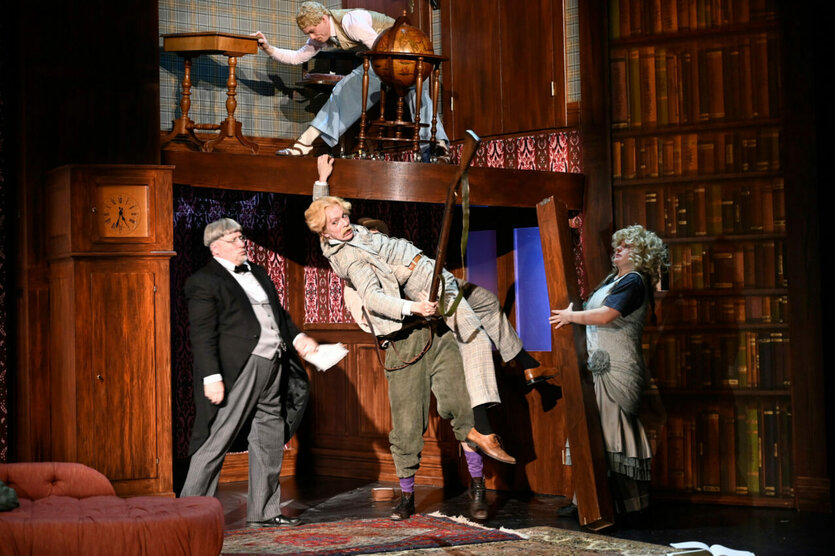 Eine Theaterbühne mit drei Schauspielern in einem Bibliothekssetting. Ein Mann wird durch eine Tür gedrückt, ein anderer steht auf einem Tisch, und eine Frau in einem Kleid beobachtet. Ein rotes Sofa und Bücherregale sind im Hintergrund.