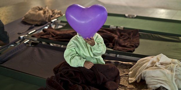 Ein Kind in einer Flüchtlingsunterkunft in Graz hält sich einen lila Luftballon in Herzform vor das Gesicht.
