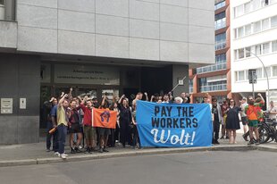 Zahlreiche Menschen stehen vor einem Gebäude und halten ein blaues Banner fest, mit der Aufschrift: Pay the Workers Wolt!