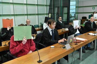 An zwei langen Tischen sitzen acht Männer, vier von ihnen halten sich Aktenordner vors Gesicht