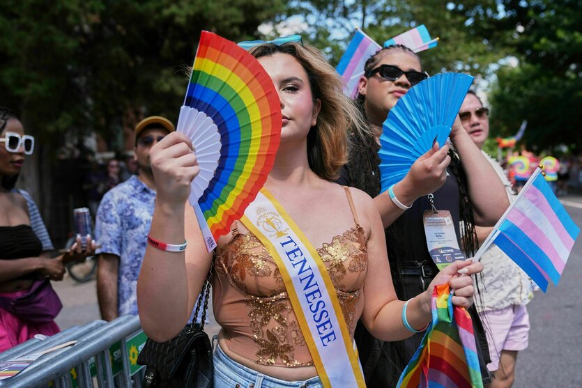 Eine Person tanzt mit anderen auf einer Straße, sie hält einen Fächer in Regenbogenfarben und Transfähnchen in den Händen, um ihren Körper trägt sie eine Scherpe auf der "Tennessee" geschrieben steht