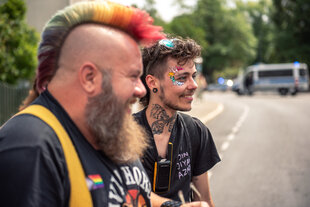 Max Armonies steht neben einer Person mit einem bunt gefärbten Iro auf einer Straße, beide lachen, im Hintergrund parkt eine Polizweiwagen