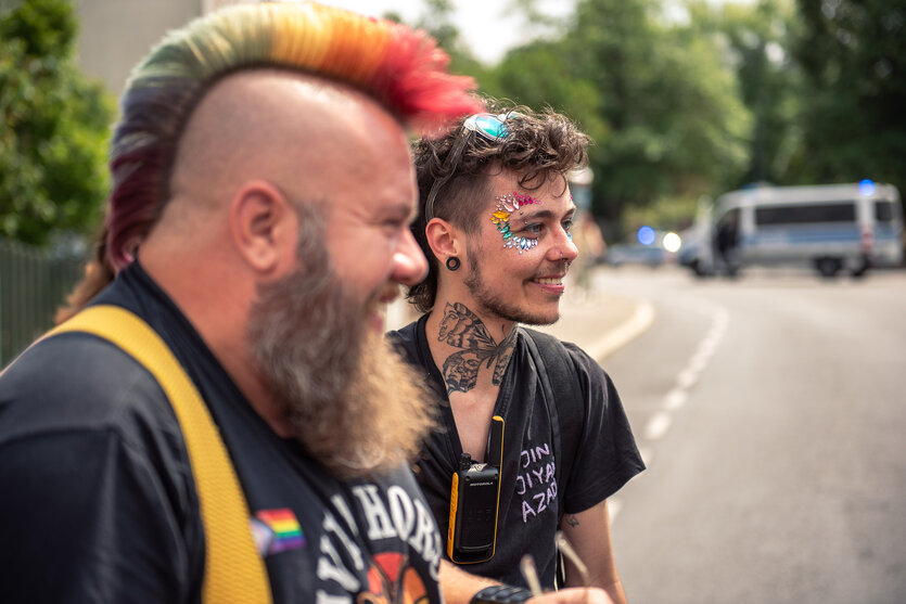 Max Armonies steht neben einer Person mit einem bunt gefärbten Iro auf einer Straße, beide lachen, im Hintergrund parkt eine Polizweiwagen