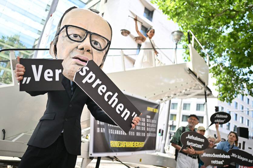 Ein Aktivist mit Merz-Maske zerbricht während einer symbolischen Protestaktion von Kabul Luftbrücke und Campact zur Unterstützung von Klagen von Afghanen gegen die Bundesregierung vor dem Auswärtigen Amt ein Schild mit der Aufschrift „Versprechen“. Im Hintergrund steht eine als Justitia verkleidete Aktivistin.