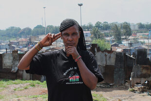 Eine Frau steht vor einer Wohnsiedlung und hält sich eine Hand unters Kinn und zwei Finger an die Schläfe. Ihr T-Shirt trägt die Aufschrift #NairobiPAMOJA.