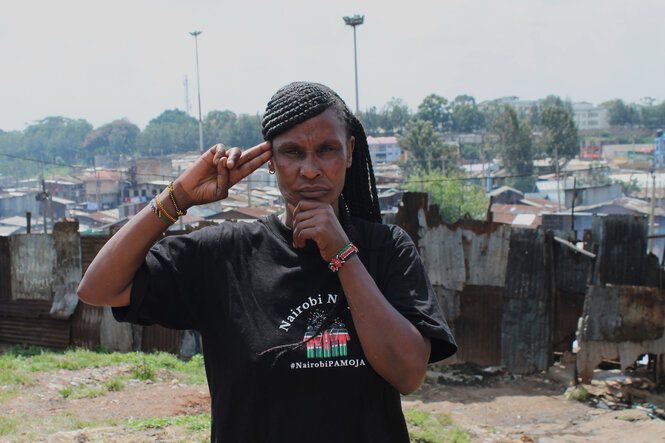 Eine Frau steht vor einer Wohnsiedlung und hält sich eine Hand unters Kinn und zwei Finger an die Schläfe. Ihr T-Shirt trägt die Aufschrift #NairobiPAMOJA.