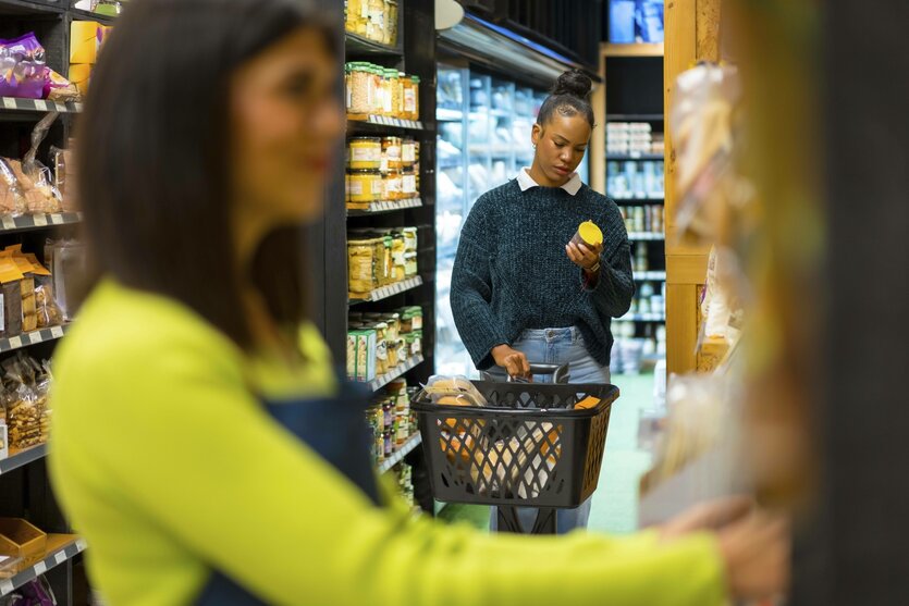 Eine Kundin wählt Bio Produkte aus, liest das Etikett und hält den Einkaufskorb im Supermarkt, während eine Verkäuferin die Regale ordnet