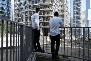 Zwei Männer stehen am Geländer eines Balkons und schauen auf von Rakenten zerstörte Hochhäuser