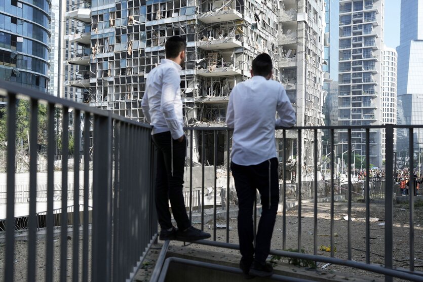 Zwei Männer stehen am Geländer eines Balkons und schauen auf von Rakenten zerstörte Hochhäuser