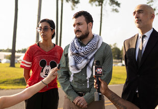 Khalil steht, an den Seiten flankiert von einer Frau und einem Mann, auf einer freien Fläche, im Hintergrund Palmen. Hände ragen ins Bild, die Khalil Mikrofone entgegenstrecken