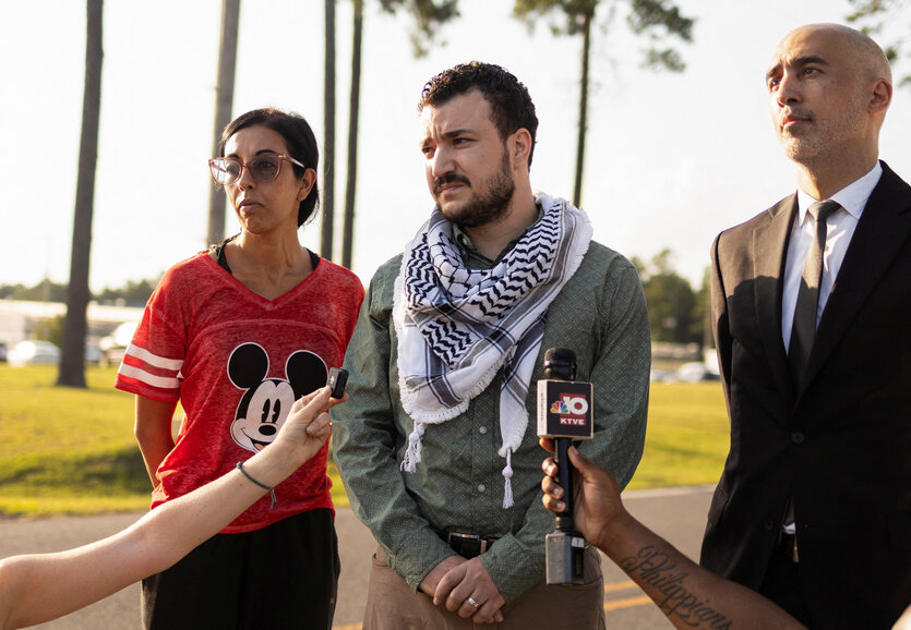 Khalil steht, an den Seiten flankiert von einer Frau und einem Mann, auf einer freien Fläche, im Hintergrund Palmen. Hände ragen ins Bild, die Khalil Mikrofone entgegenstrecken