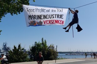 Aktivist hängt an einem Seil hängend ein Transparent mit der Aufschrfit „Organisiert euch gegen Klimakrise und Aufrüstung“ auf