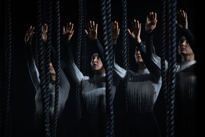 Vier Theaterdarstellerinnen mit ernsten Gesichtern und erhobenen Händen auf einer dunklen Theaterbühne