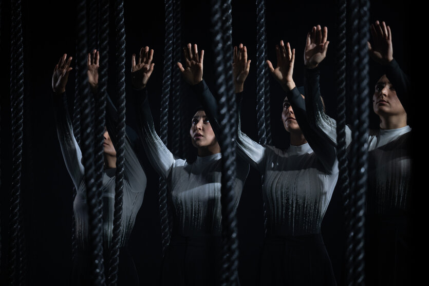 Vier Theaterdarstellerinnen mit ernsten Gesichtern und erhobenen Händen auf einer dunklen Theaterbühne