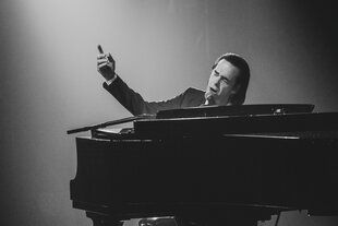 Nick Cave, singend an einem Flügel sitzend, die rechte Hand gehoben, der Zeigefinger zeigt nach oben links, von wo Licht auf ihn fällt