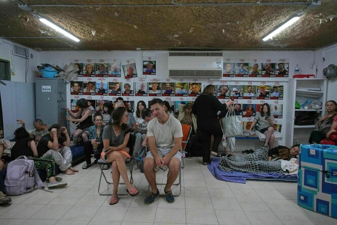 Ein Mann und eine Frau sitzen auf Klappstühlen, im Hintergrund hocken Menschen auf dem Boden, an der Wand die Fotos von Menschen, die als Geiseln nach Gaza verschleppt wurden