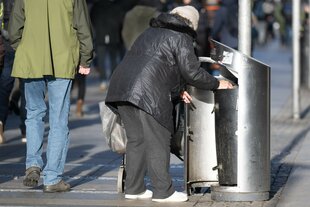 Zu sehen ist eine Person die in einem Mülleimer nach Pfandflaschen sucht. Ihre Körperhaltung deutet darauf hin, dass es sich um eine Person fortgeschrittenen Alters handelt.