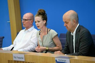 Drei Personen sitzen nebeneinander und sprechen zu Vertretern der Presse
