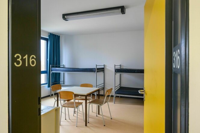 Blick durch eine Tür in einen kargen Raum mit Hochbetten und einem Tisch mit Stühlen