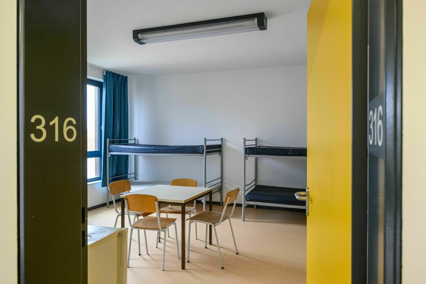 Blick durch eine Tür in einen kargen Raum mit Hochbetten und einem Tisch mit Stühlen