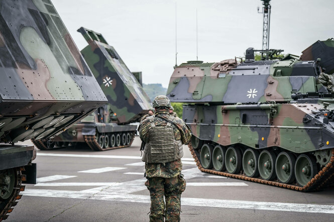 Ein Soldat in Gefechtsausrüstung steht zwischenMehrfachraketenwerfer und Panzer