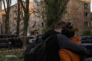 Im Vordergrund zwei Menschen, die sich umarmen, im Hintergrund ein zerstörtes Gebäude
