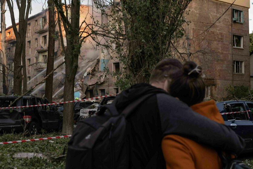 Im Vordergrund zwei Menschen, die sich umarmen, im Hintergrund ein zerstörtes Gebäude