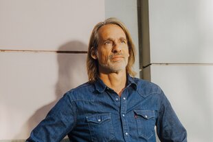 Zu sehen ist Richard David Precht vor einem weißen Hintergrund. Er trägt ein blaues Denim-Hemd, wird von der Sonne angeschienen und schaut in die Ferne.