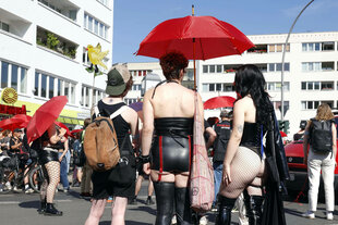Demo, Plattenbauten, drei Personen die uns den Rücken zu weden sind unter einem roten Schirm bei strahelendem Sonnenschein. Sie Tragen freizügige fetish Kleidung.