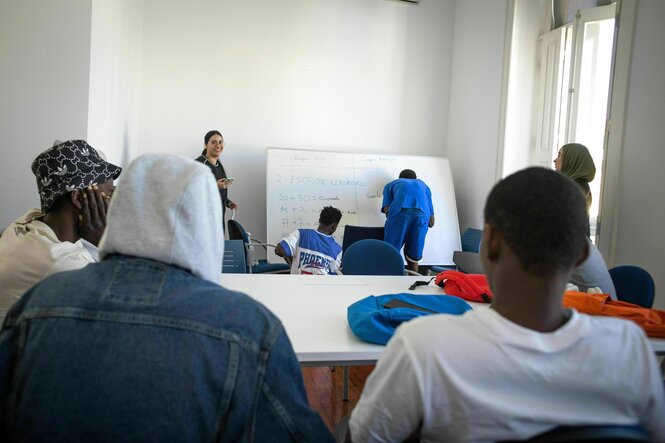 Blick in ein Klassenzimmer, die jungen Männer schauen auf zwie Klassenkameraden die an die Tafel schreiben, die Lehrerin steht freundlich lächelnd neben ihnen