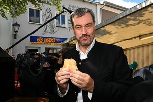 Der bayerische Ministerpräsident Markus Söder posiert mit einer Fleischsemmel neben einem Grillmeister